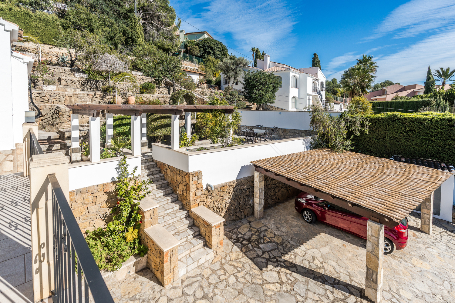 Vista exterior con entrada empedrada, aparcamiento cubierto con vigas de madera y jardín mediterráneo aterrazado con muros de piedra natural.