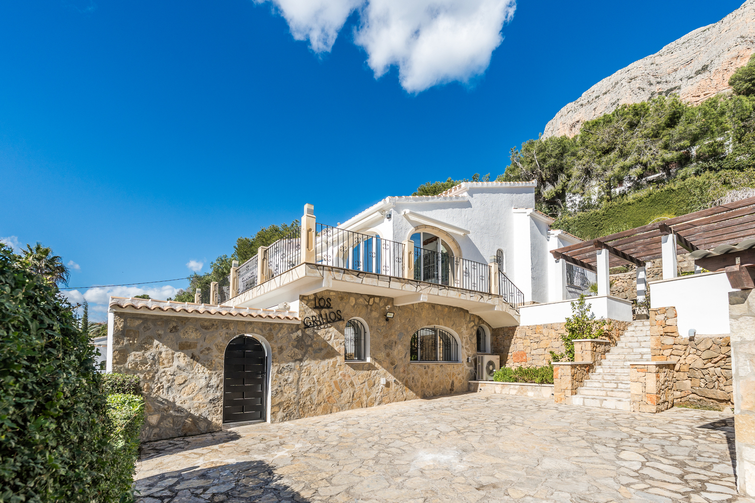 Vista exterior con fachada de piedra natural, ventanas de arco y amplia zona de aparcamiento pavimentada bajo el macizo del Montgó.