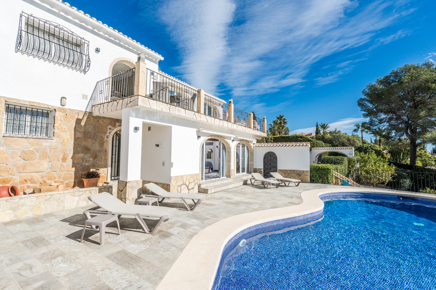 Vista exterior de la piscina de mosaico curvo y terraza de piedra, con tumbonas blancas y fachada de la villa con zócalo de piedra natural y arcos.