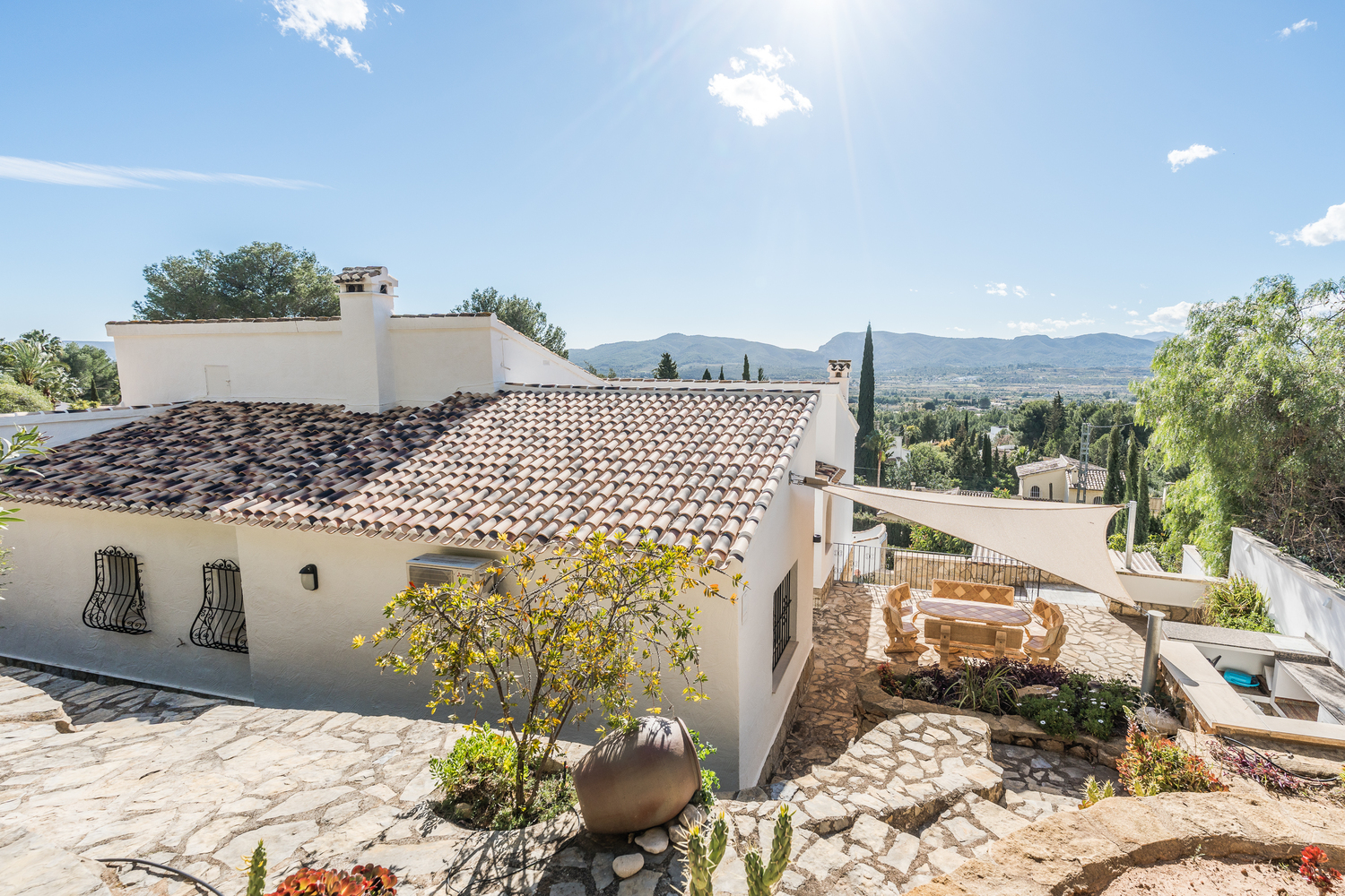 Vista exterior de la villa con tejados de terracota, terrazas de piedra natural, zona de comedor bajo un toldo vela y vistas panorámicas al valle y las montañas.