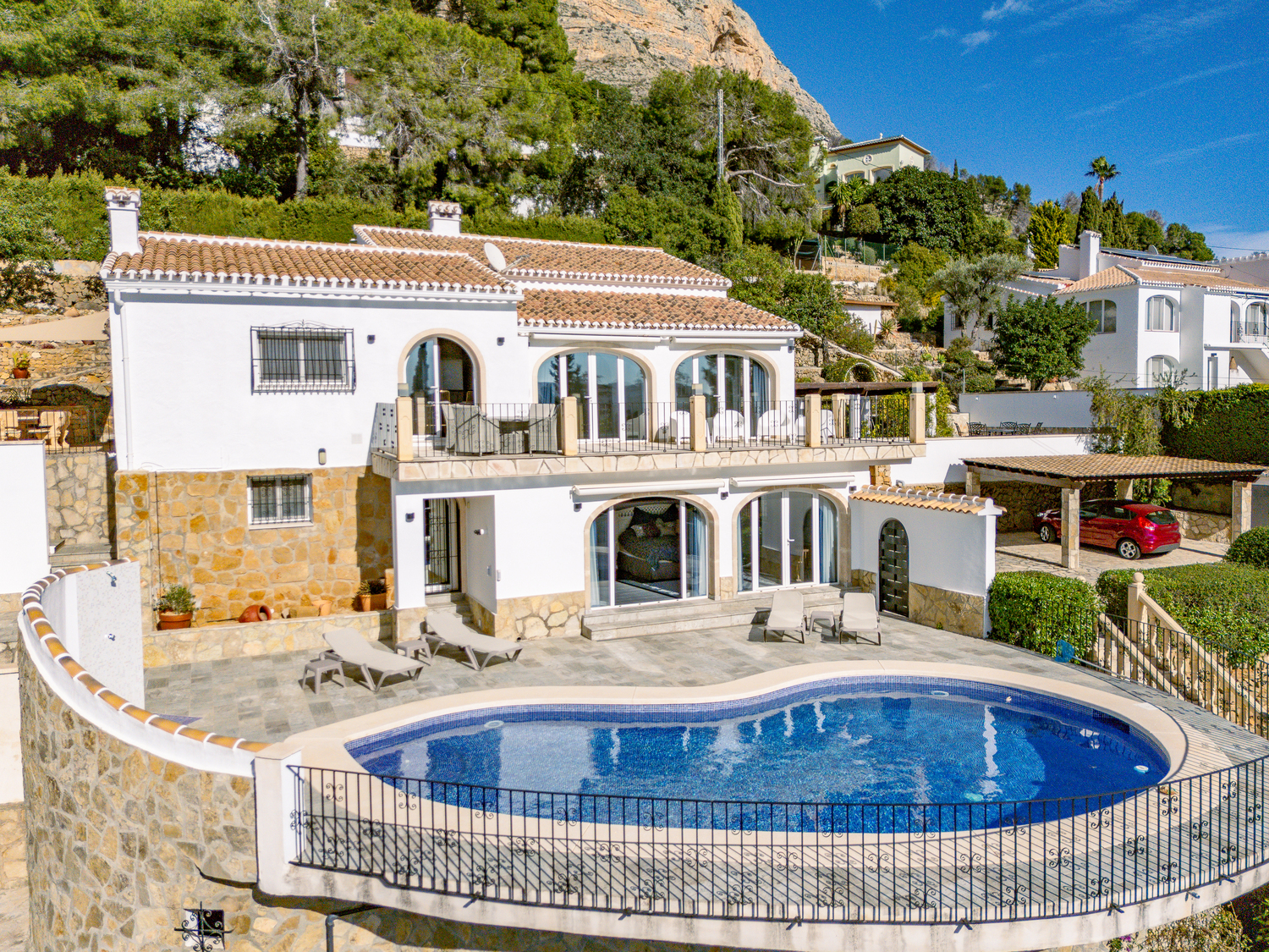 Vista exterior de la villa de dos niveles con piscina curva, terraza de piedra y ventanales en arco, situada bajo el macizo del Montgó en Jávea.