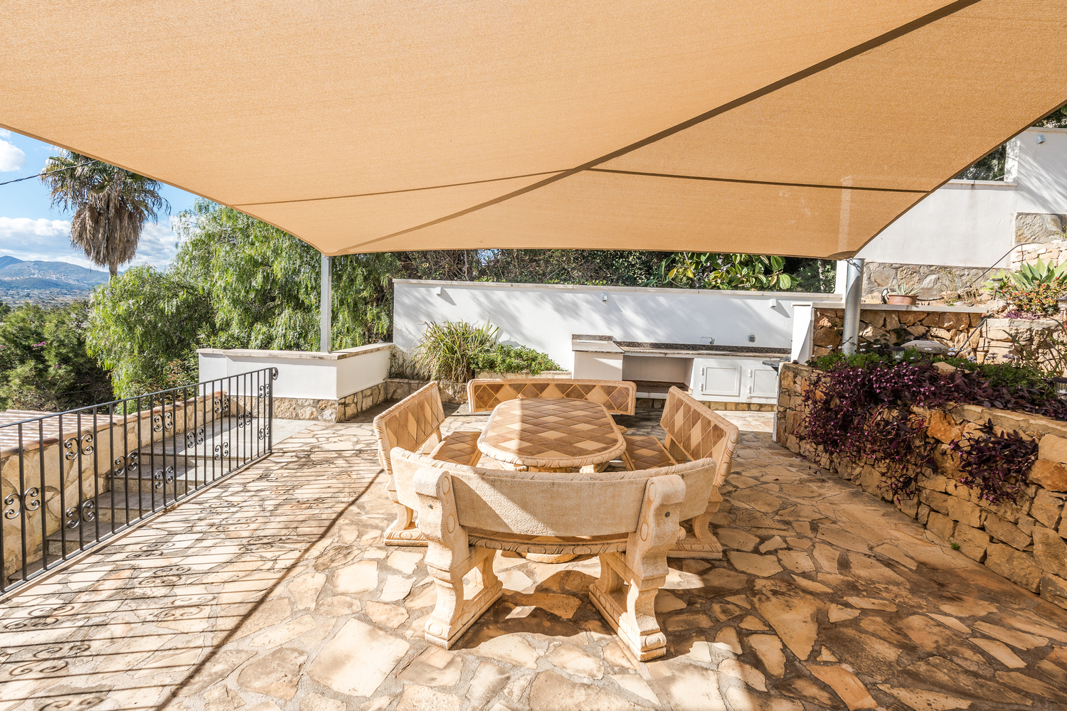 Zona de comedor exterior con mesa y bancos de piedra tallada, protegida por un toldo vela sobre una terraza de piedra natural junto a la cocina de verano.