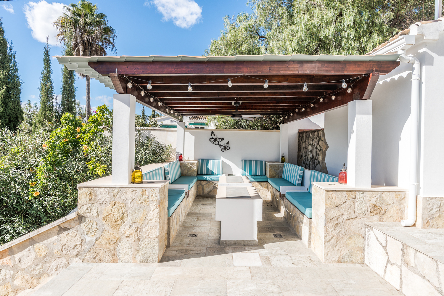 Zona de estar exterior cubierta con pérgola de madera, ventilador de techo y bancos de piedra integrados con cojinería a rayas turquesas y blancas.