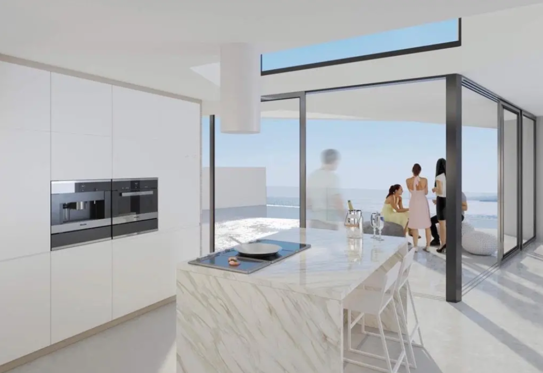 Cocina moderna con isla de mármol y placa integrada, armarios minimalistas blancos y amplios ventanales que conectan con la terraza y vistas al mar.