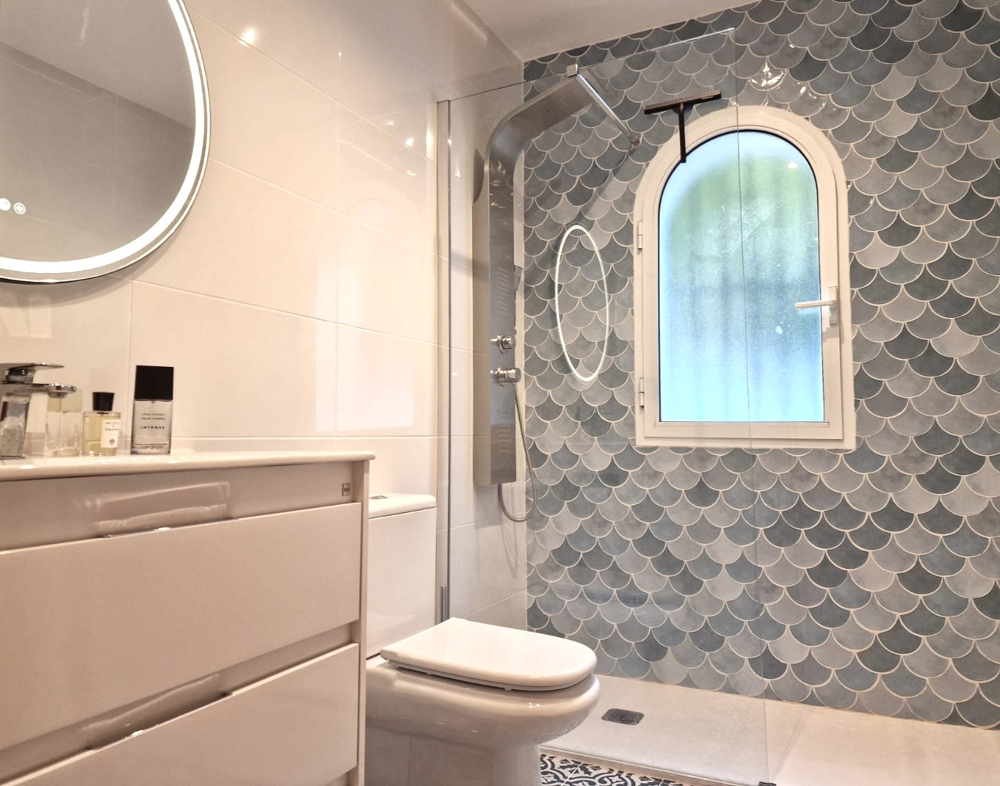 Baño moderno con ducha a ras de suelo, azulejos de escama azules, ventana de arco con cristal translúcido, columna de ducha de acero y espejo circular retroiluminado.
