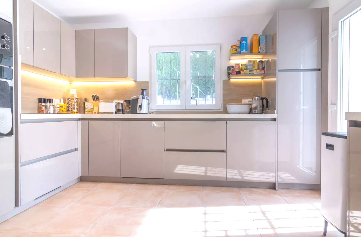 Cocina moderna con armarios en color taupe brillante, encimeras blancas e iluminación LED integrada. Incluye electrodomésticos encastrados y suelo de baldosas cerámicas.