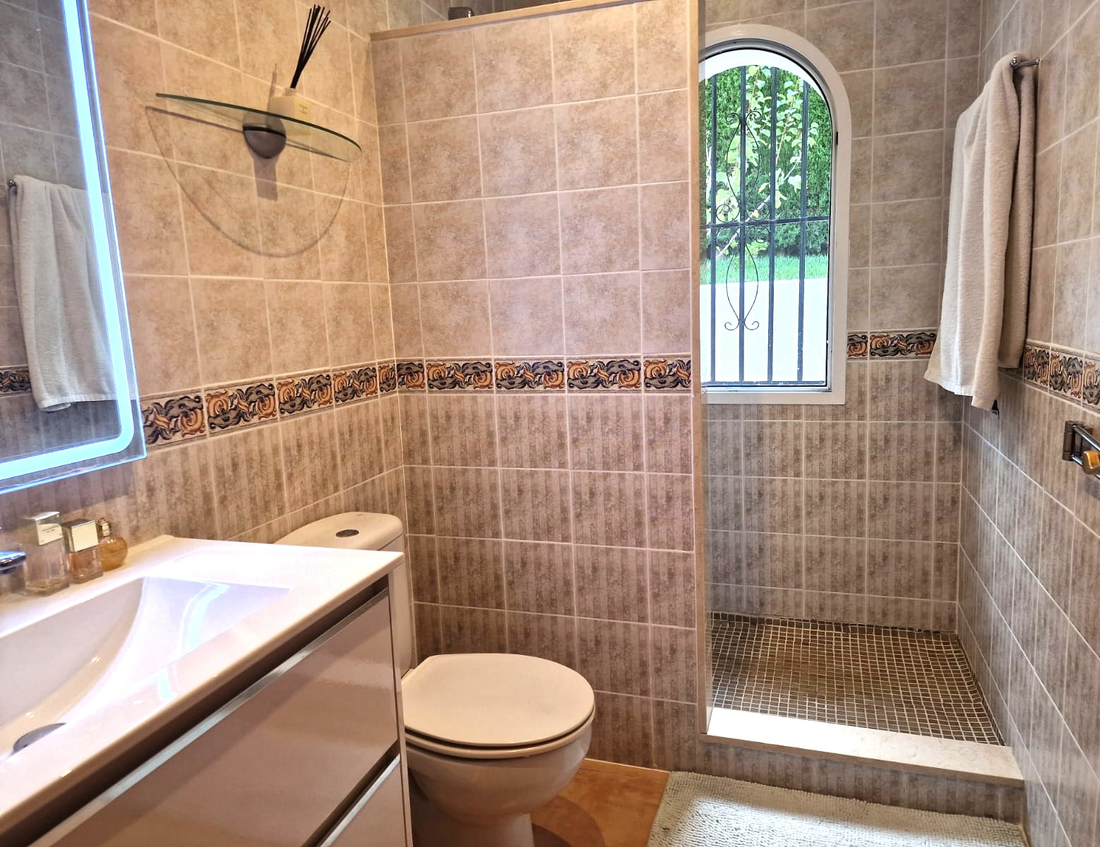 Cuarto de baño moderno con ducha a ras de suelo, paredes alicatadas en tonos beige con cenefa decorativa y una ventana de arco que aporta luz natural.