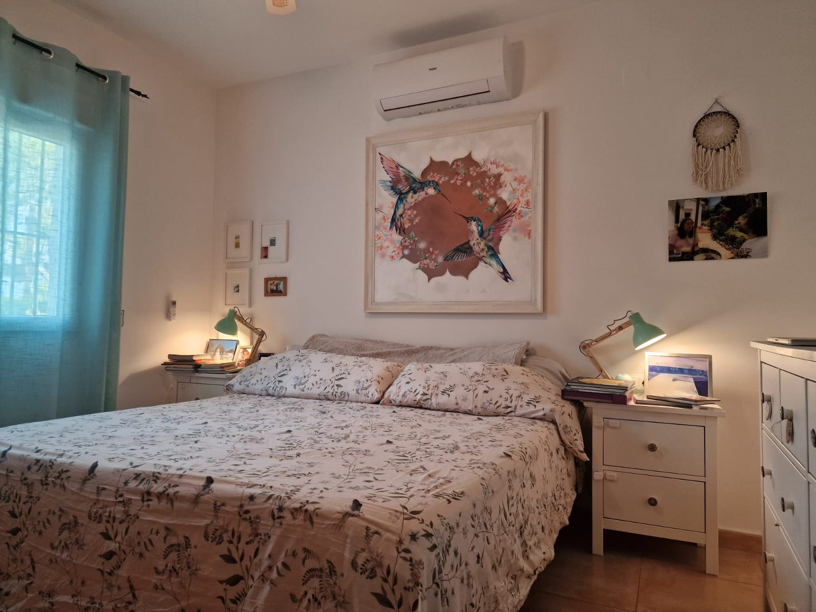 Dormitorio luminoso con cama doble, aire acondicionado Haier, mesitas de noche con lámparas flexo verdes y un gran cuadro decorativo de colibríes sobre el cabecero.