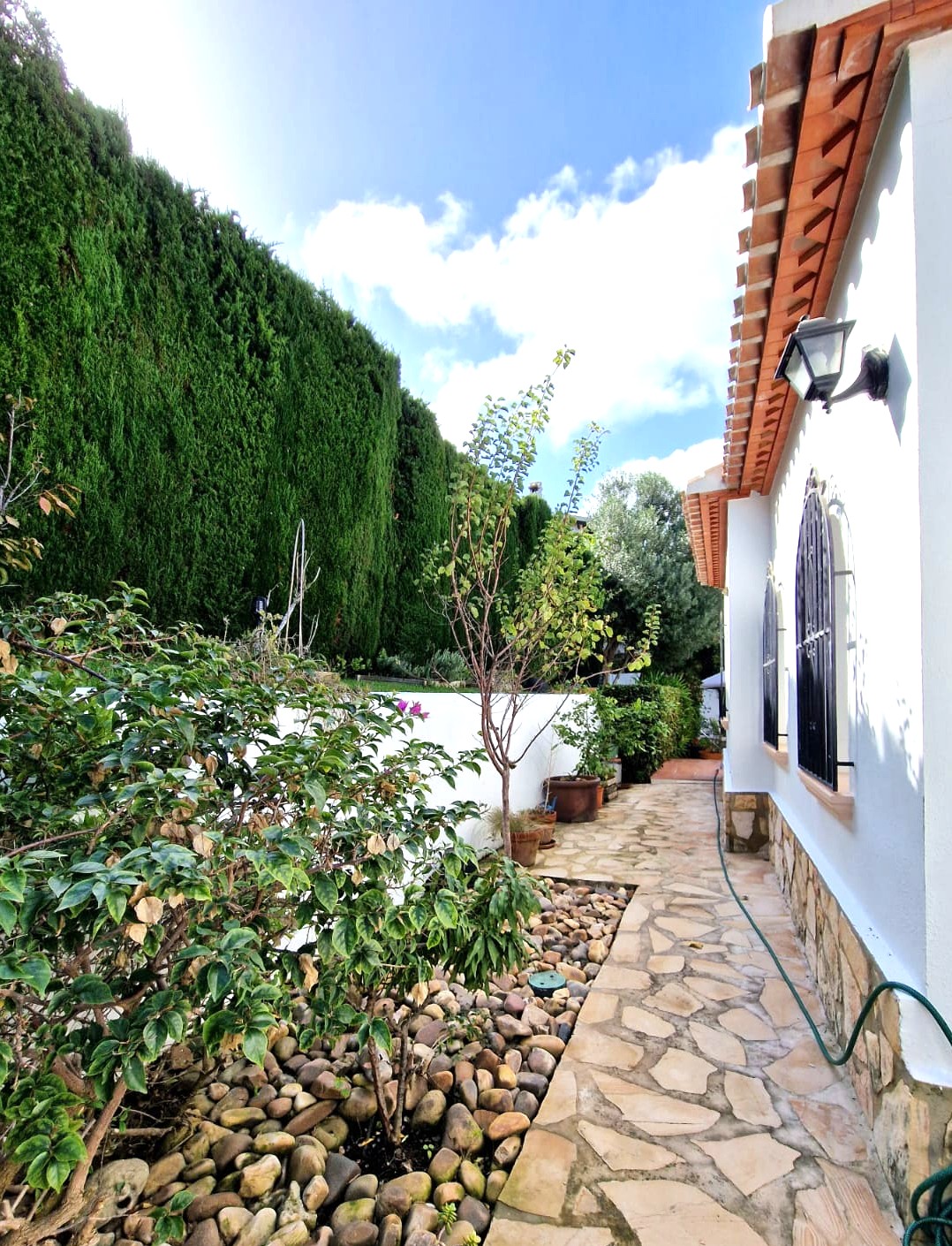 Pasillo lateral con pavimentación de piedra natural, fachada blanca con zócalo de piedra y setos de ciprés. Ventanas con rejas de hierro y tejas cerámicas tradicionales.