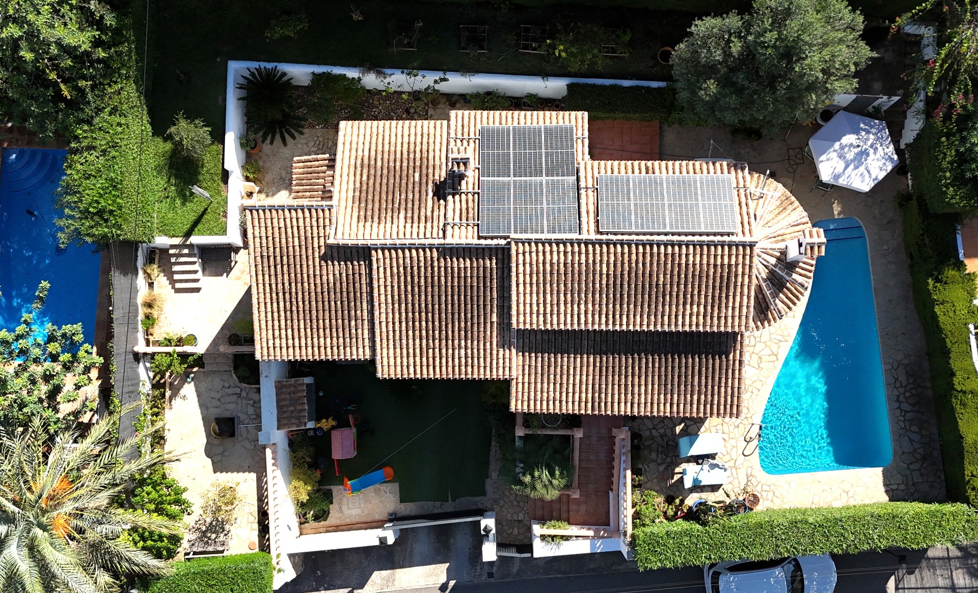 Perspectiva aérea de la villa que destaca el tejado de tejas con paneles solares, la piscina privada y un jardín mediterráneo con zona de juegos infantil.