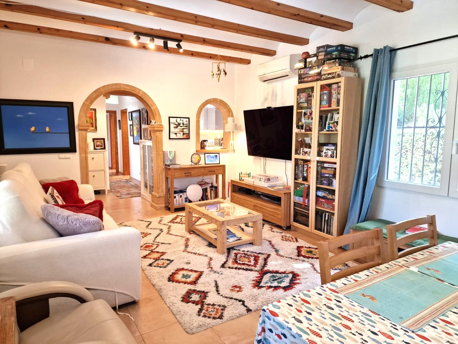 Salón comedor con vigas de madera vistas, arcos de piedra Tosca y aire acondicionado. El espacio integra una zona de estar luminosa con acceso al pasillo y la cocina.