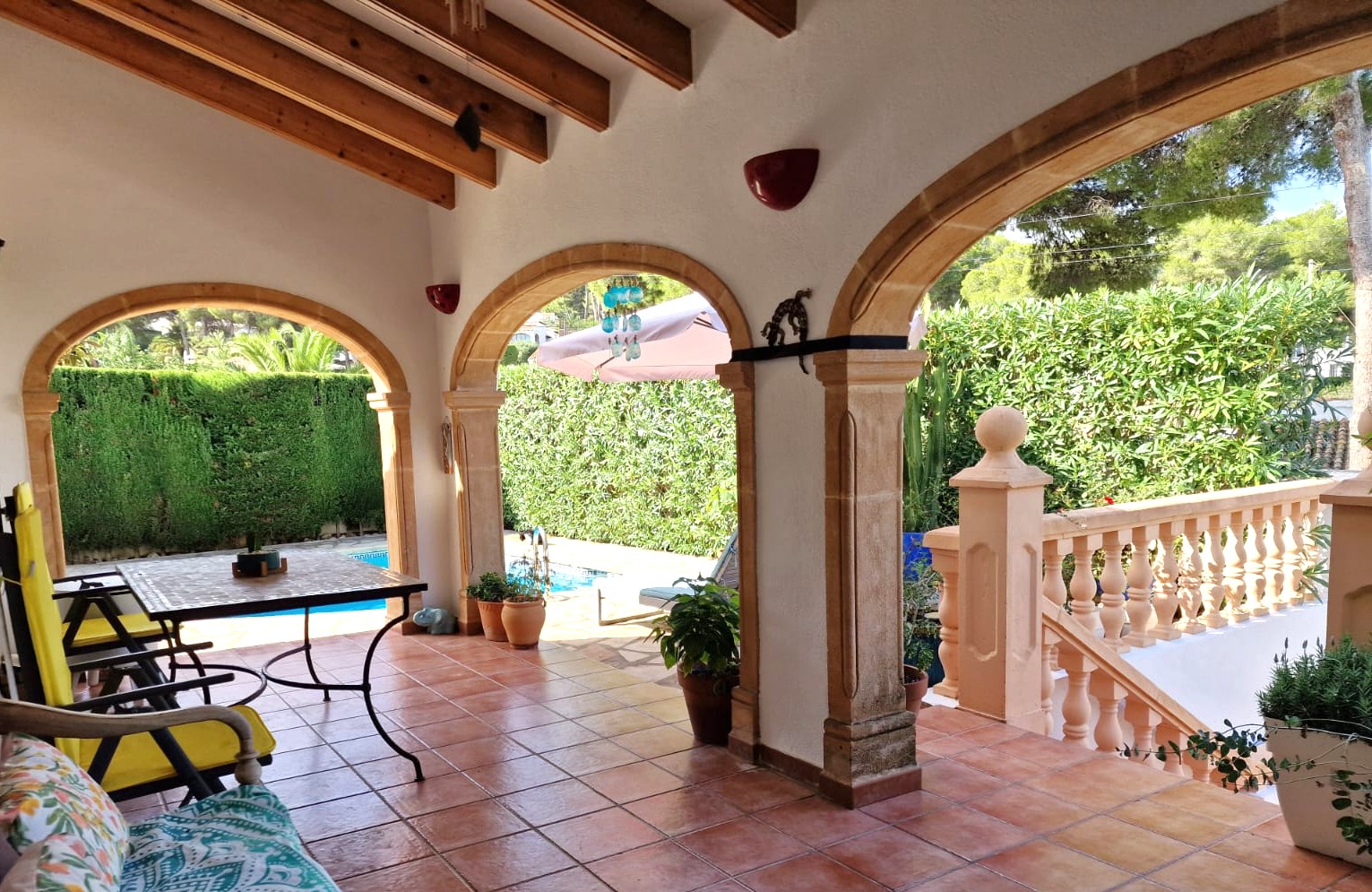 Terraza cubierta con arcos de piedra tosca, vigas de madera a la vista y suelo de barro, con vistas directas a la piscina y al jardín privado.