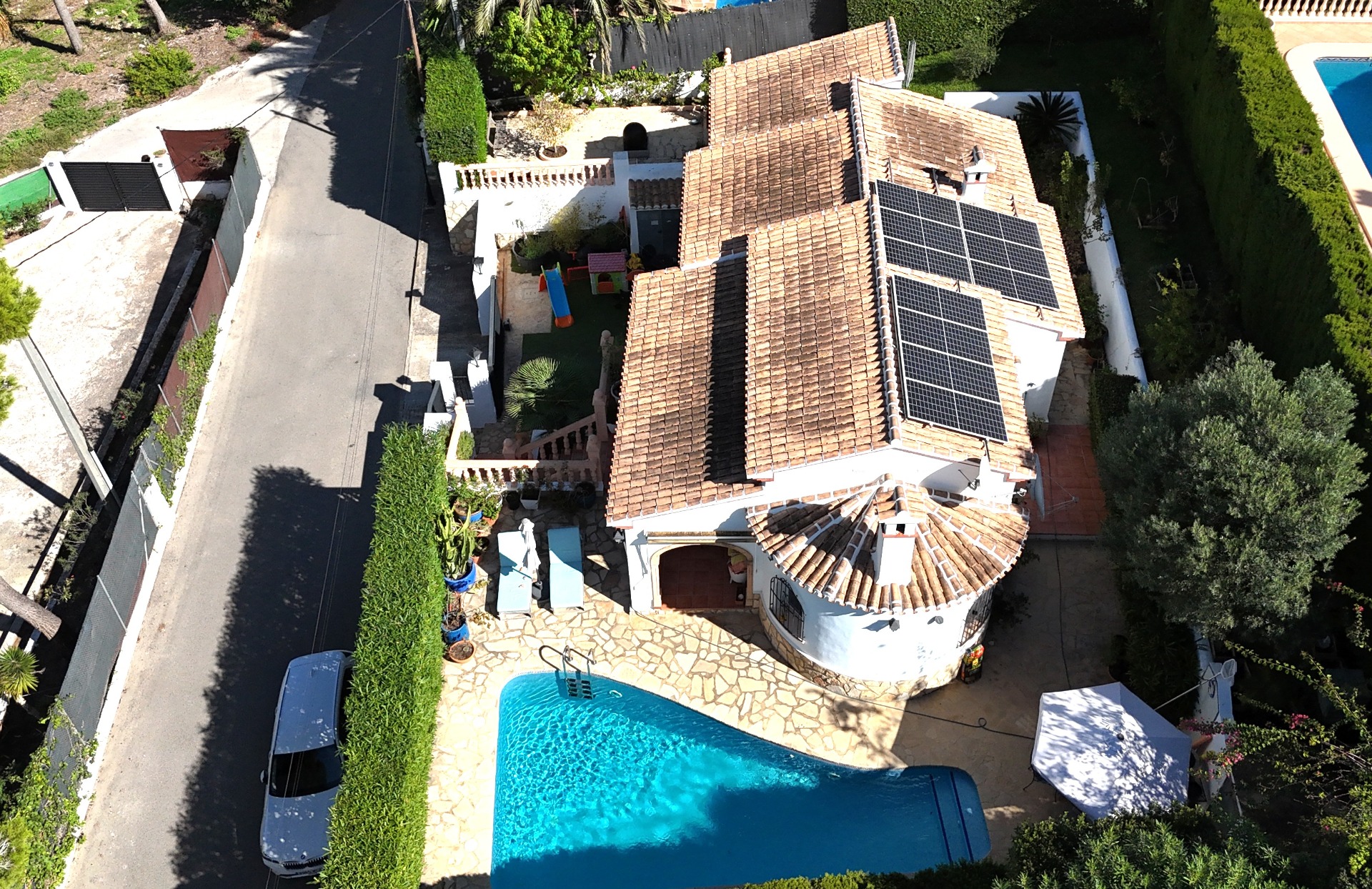 Vista aérea de la villa que muestra la piscina privada, la instalación de paneles solares sobre el tejado de teja y una zona de juegos infantiles en la parcela vallada.