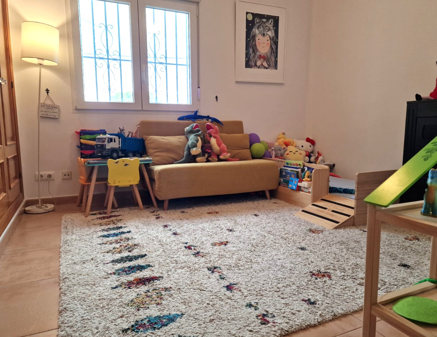 Zona de juegos infantil con alfombra de pelo largo, sofá amarillo y mesa de actividades. La estancia cuenta con luz natural, suelos cerámicos y ventana con rejas decorativas.