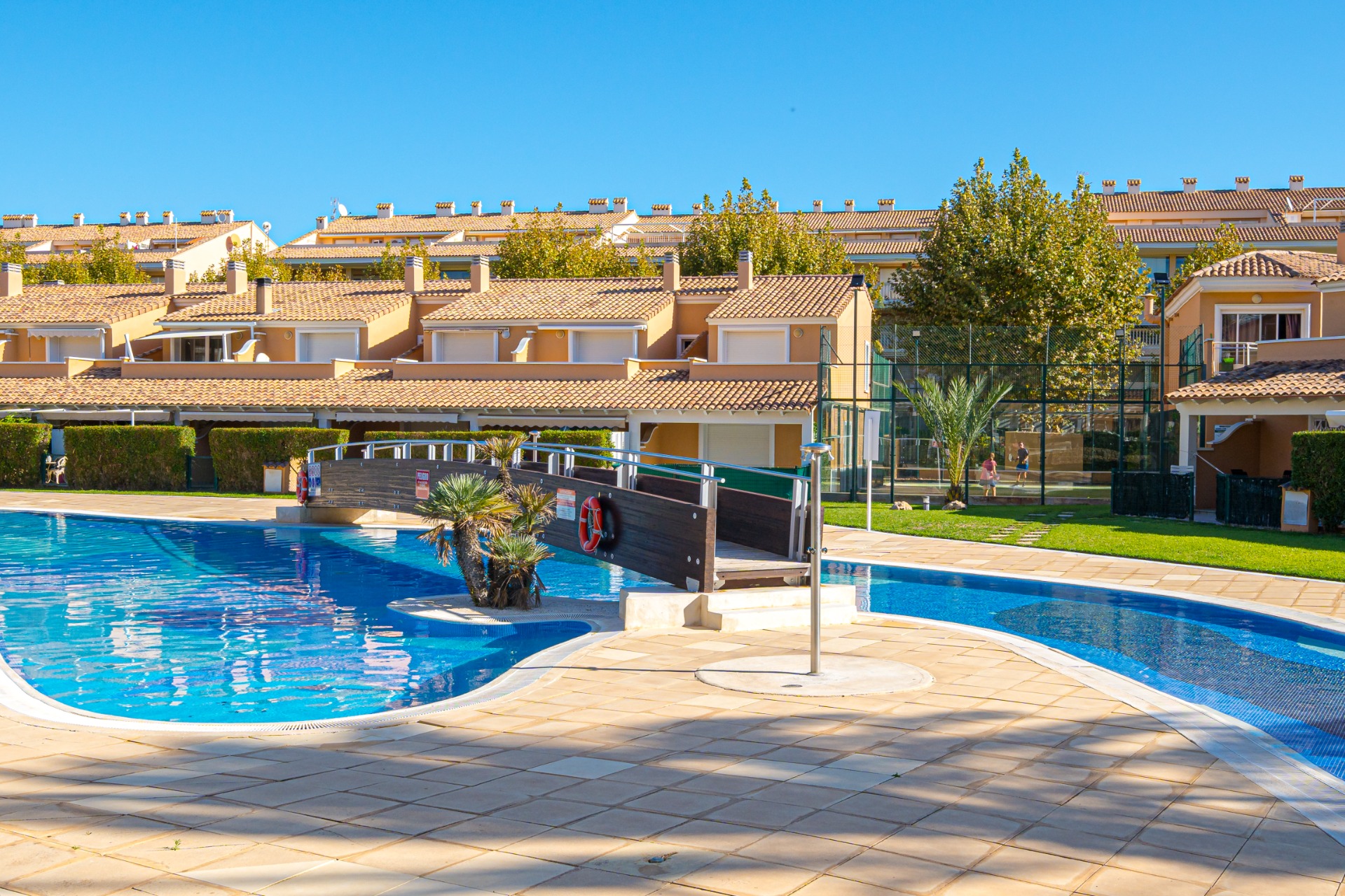 Amplia piscina comunitaria con puente de madera, rodeada de una terraza solárium y pistas de pádel al fondo bajo el sol del Mediterráneo.
