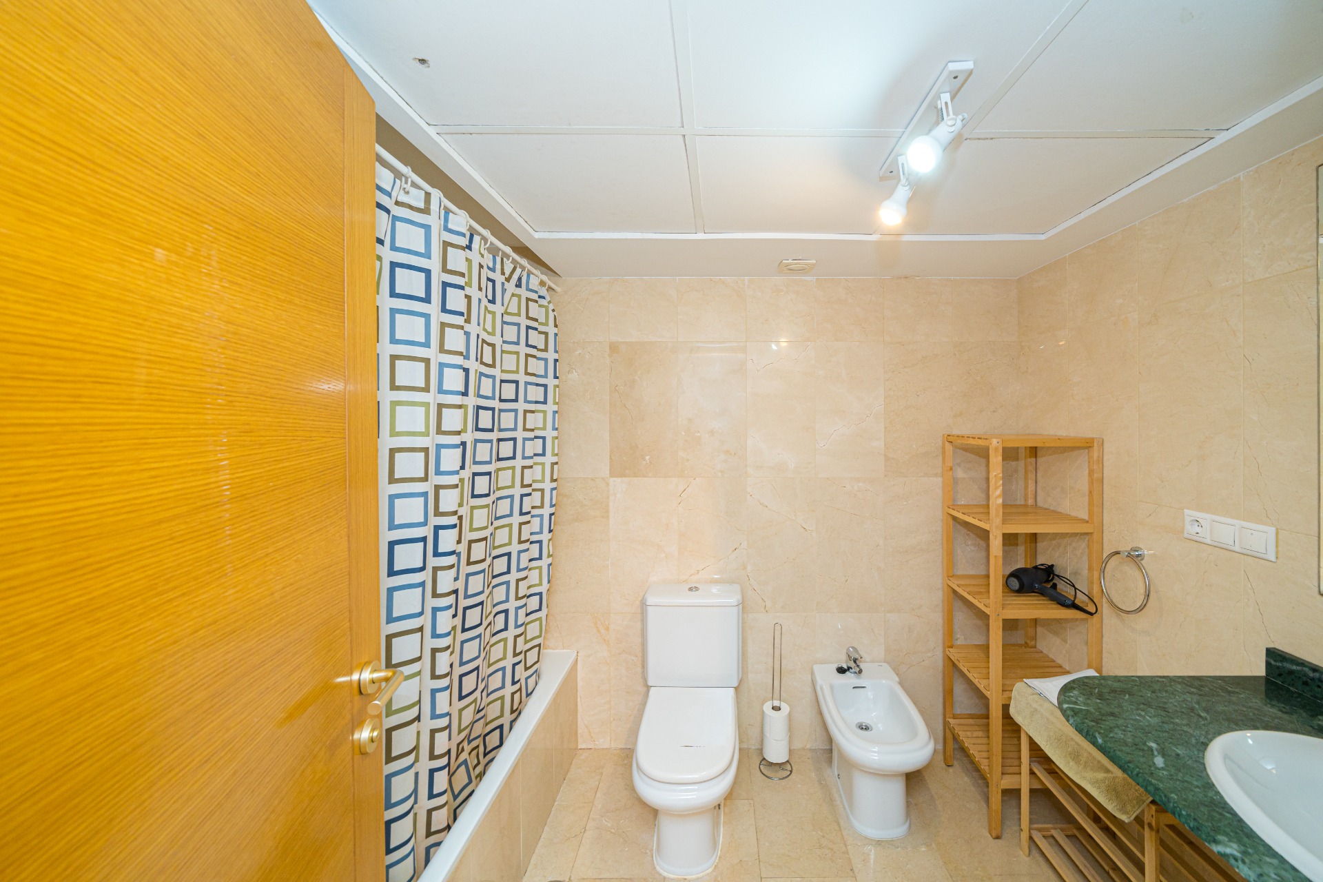 Cuarto de baño con revestimiento de mármol beige, equipado con bañera, bidé y una encimera de mármol verde sobre una estructura de madera funcional.