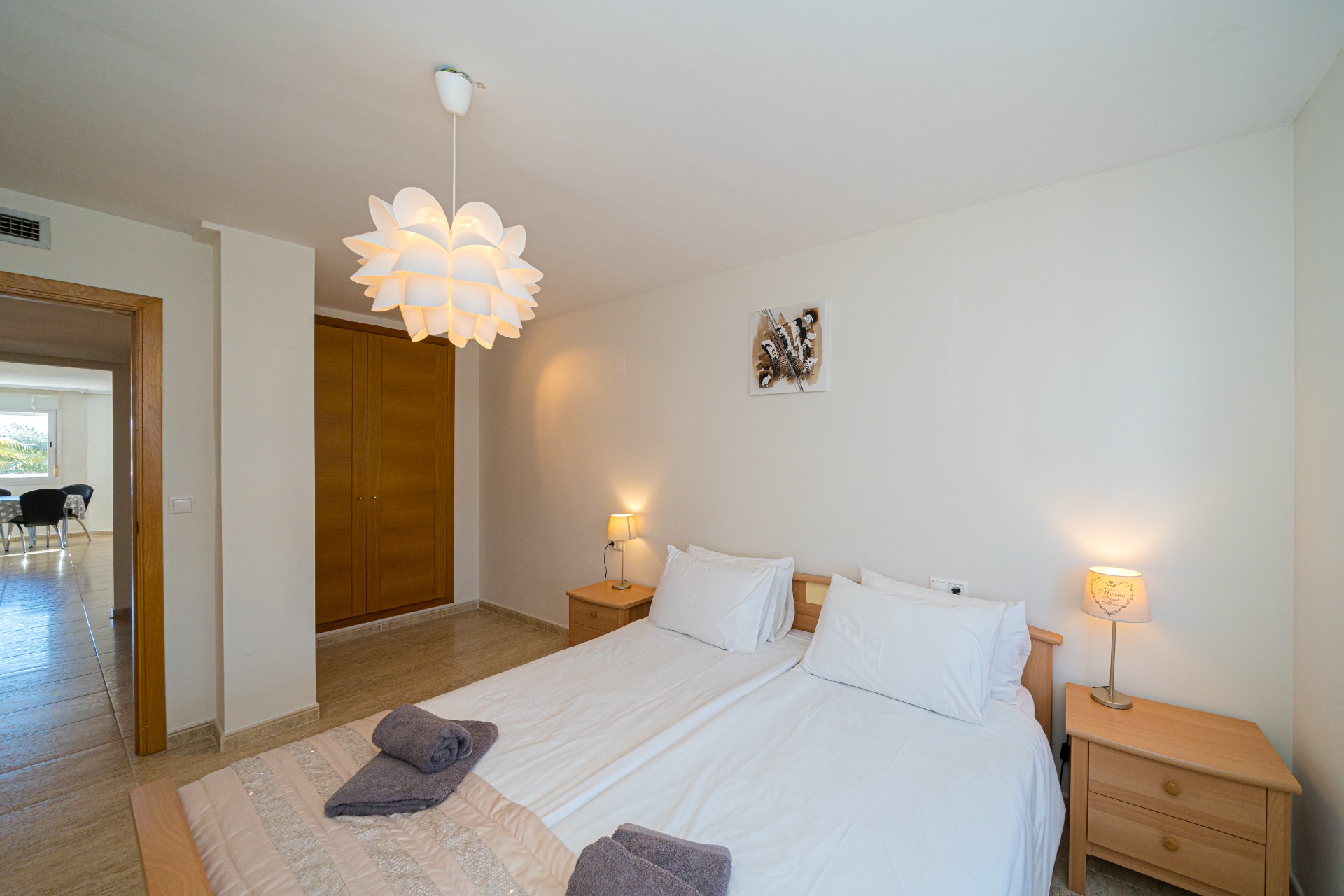 Dormitorio con camas dobles, armarios empotrados de madera y una lámpara de techo decorativa. La vista a través de la puerta muestra un comedor luminoso.