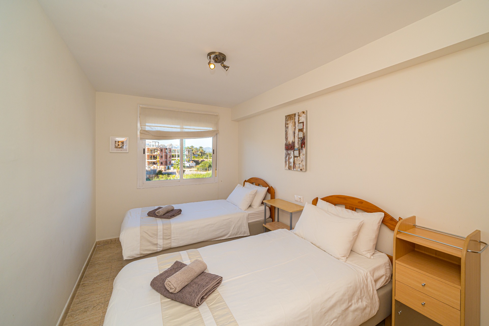Dormitorio luminoso con dos camas individuales, cabeceros de madera, suelos de gres y una ventana que ofrece luz natural y vistas despejadas al exterior.
