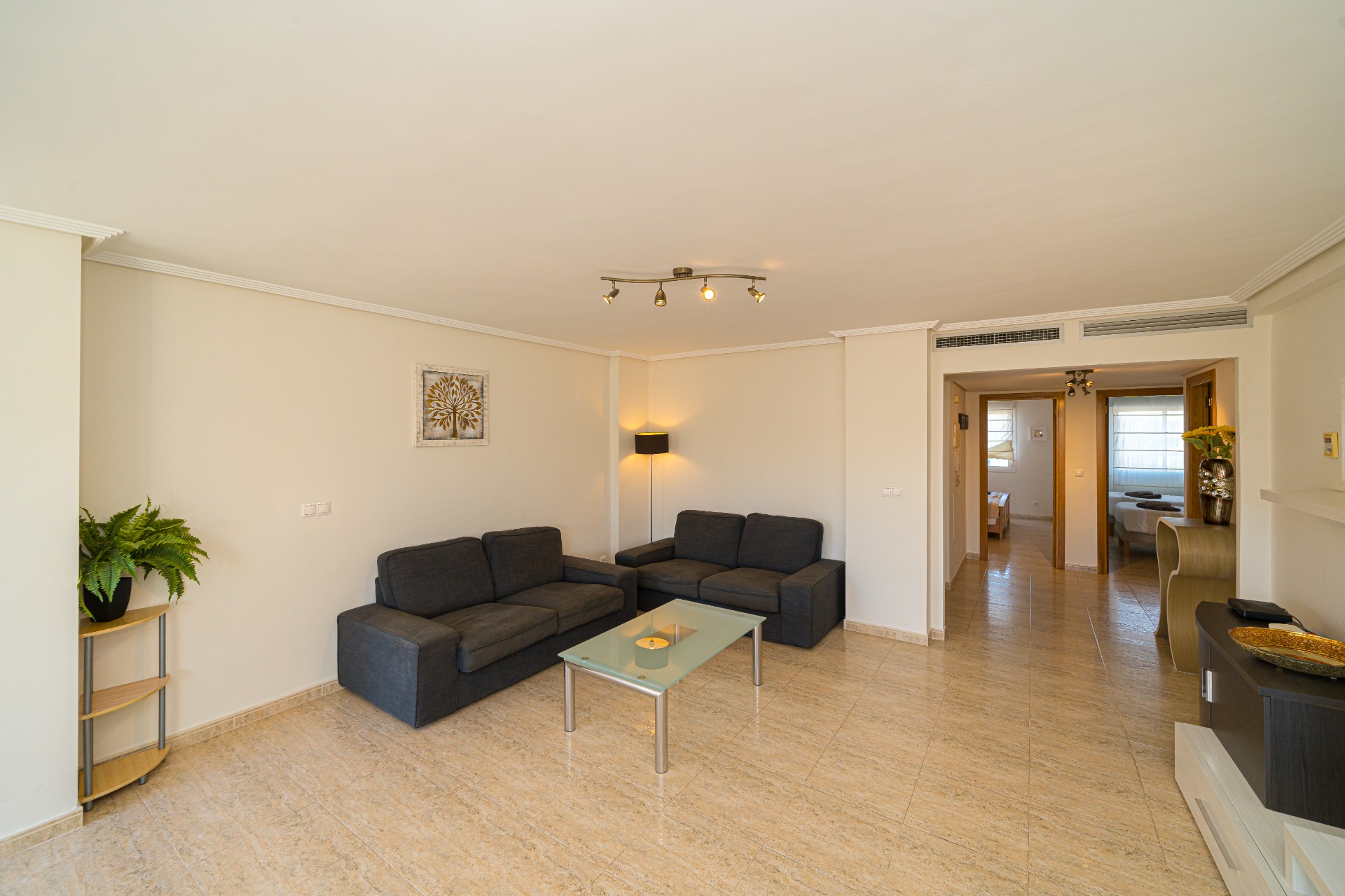Salón espacioso con sofás en gris oscuro, mesa de centro de cristal y suelos de gres pulido. La distribución abierta conecta directamente con el pasillo de los dormitorios.