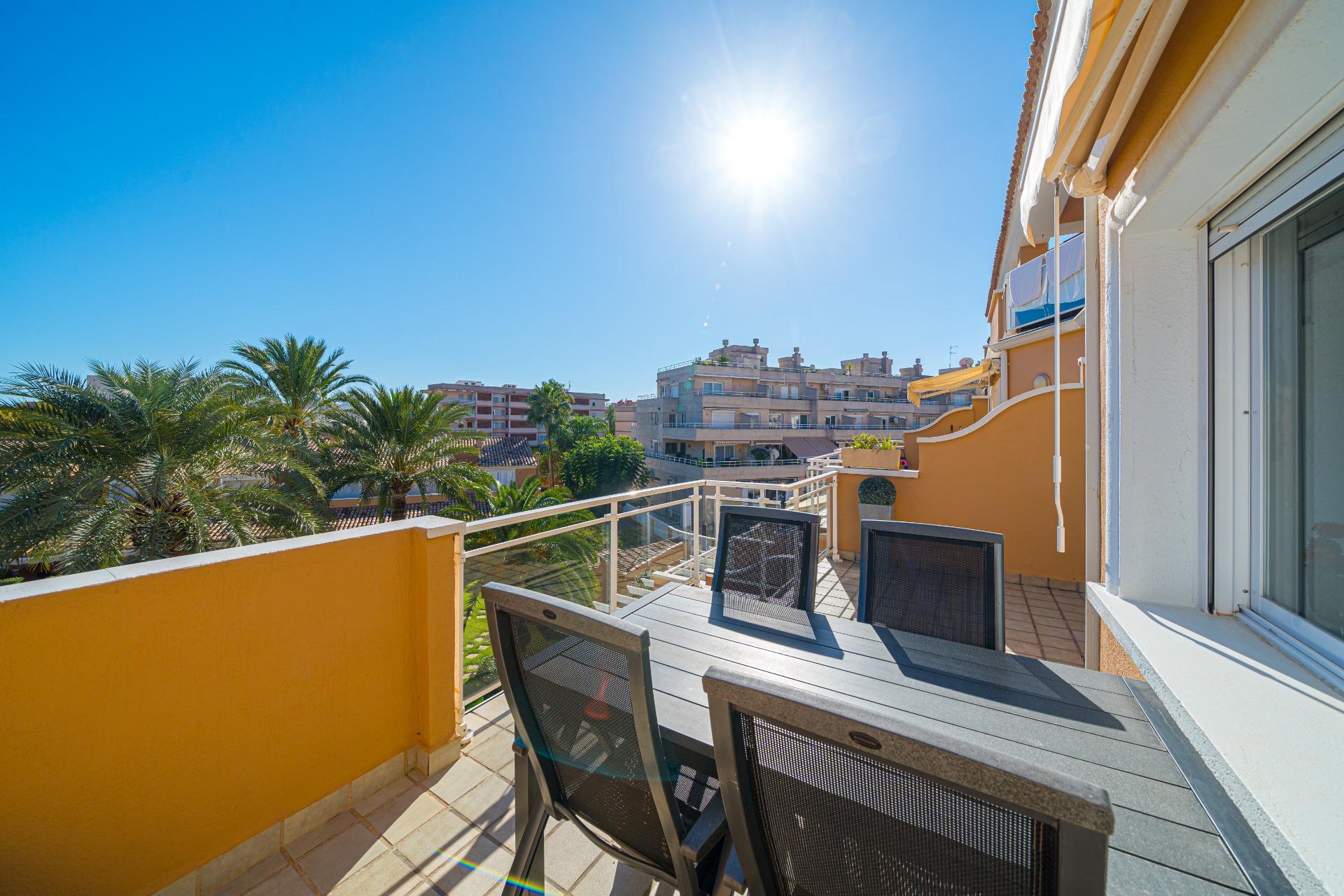 Terraza privada soleada con conjunto de comedor exterior en gris oscuro, suelo cerámico y vistas despejadas a las palmeras y complejos residenciales.