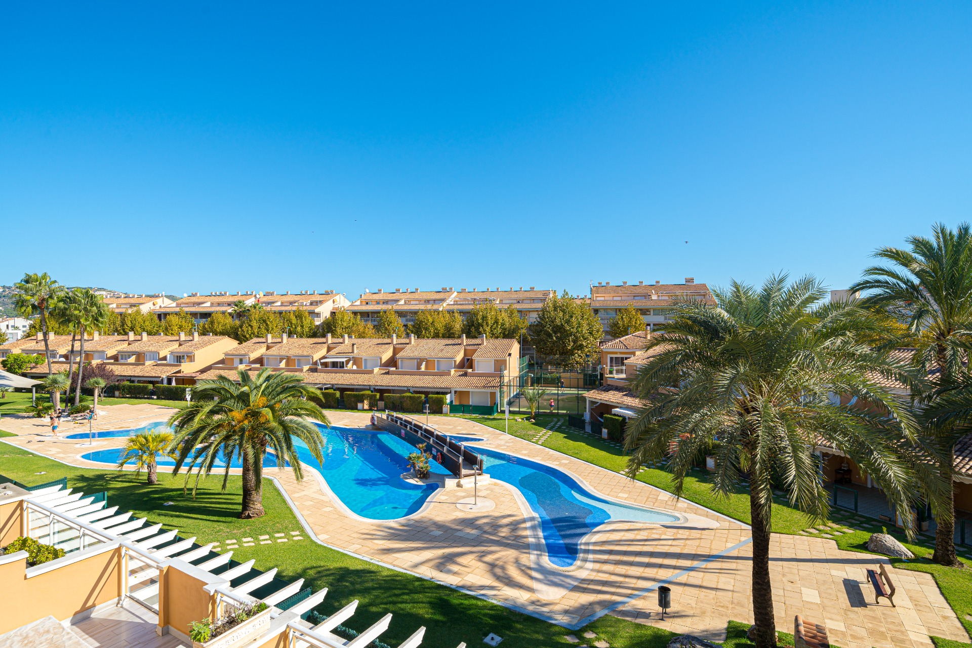 Vista elevada de la piscina comunitaria con puente de madera, rodeada de jardines cuidados, palmeras y la pista de pádel del complejo.