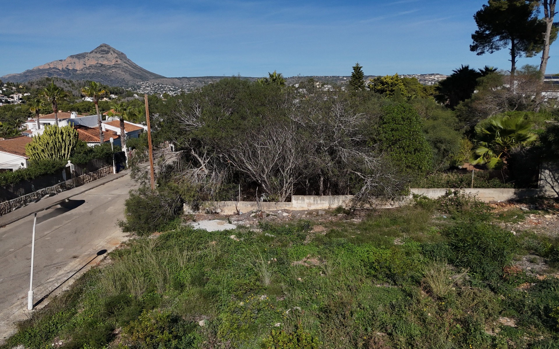 Vista elevada de la parcela llana de 889 m² en Pinosol, destacando el acceso por calle residencial, el muro perimetral y las vistas despejadas al macizo del Montgó.