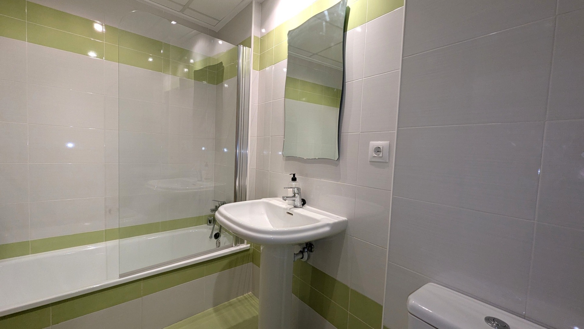 Baño funcional con bañera y mampara de cristal, lavabo de pedestal y revestimiento cerámico en blanco y verde lima con espejo de diseño ondulado.