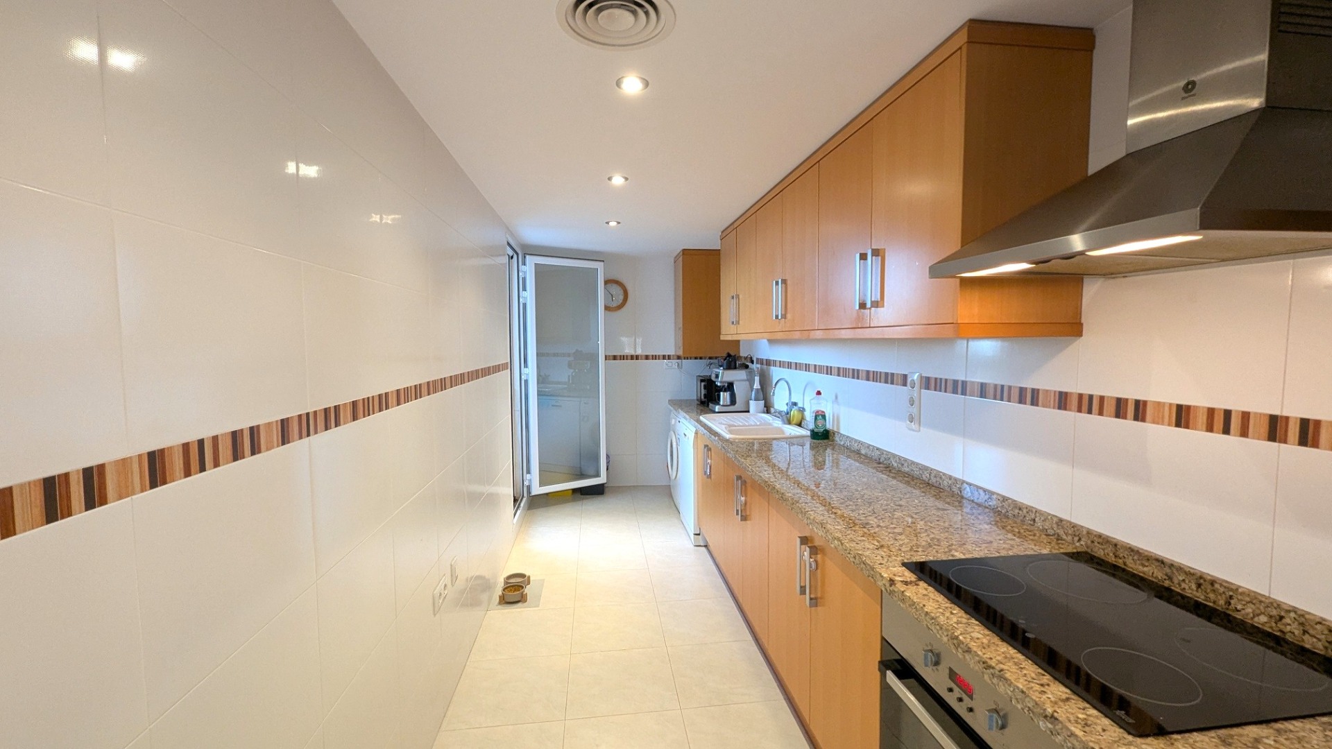 Cocina independiente con armarios de madera, encimera de granito y electrodomésticos integrados. Incluye paredes alicatadas en blanco con cenefa decorativa e iluminación empotrada.