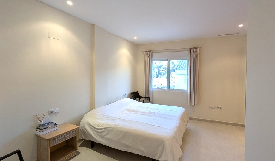 Dormitorio luminoso con tonos neutros, focos empotrados en el techo y ventana con persianas. Dispone de suelos cerámicos y una mesita de noche de madera.