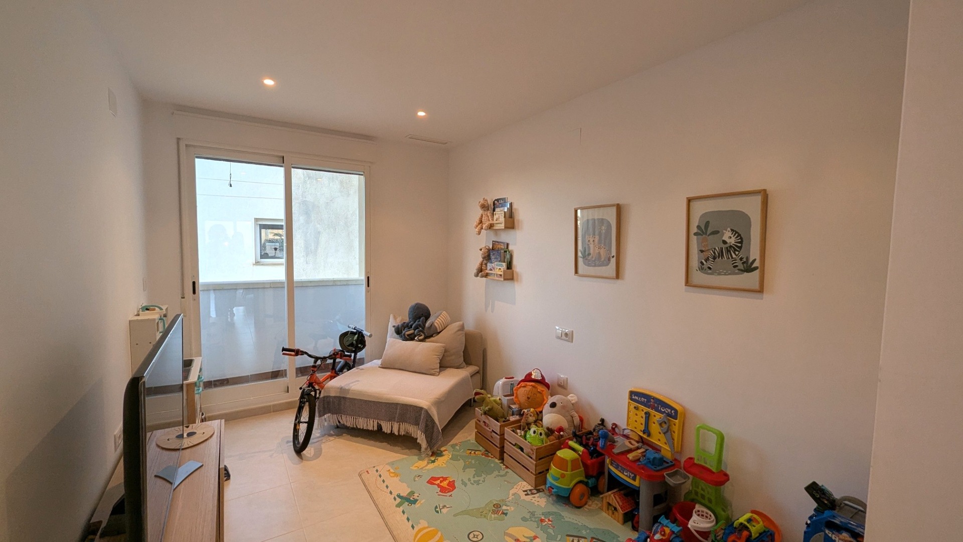 Dormitorio luminoso utilizado como cuarto de juegos, con focos empotrados, suelos de gres claro y acceso directo a la terraza mediante puertas correderas.