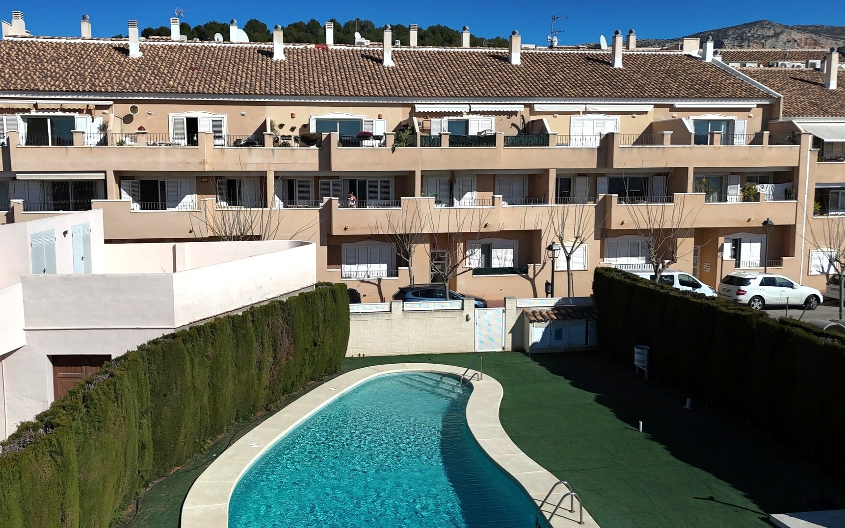 Vista de la piscina comunitaria de forma arriñonada rodeada de césped artificial y setos altos, frente a la fachada del complejo con persianas blancas.