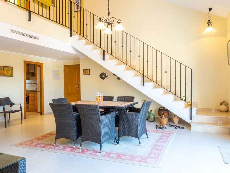 Zona de comedor con sillas de mimbre oscuro bajo una escalera de tonos claros y barandilla de hierro forjado. Espacio abierto con suelos cerámicos y luz natural.