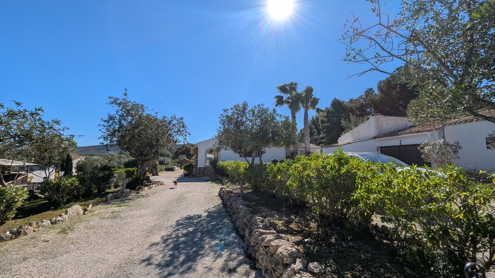 Amplio camino de grava flanqueado por muros de piedra seca que conduce a la vivienda principal de estilo finca, rodeada de vegetación mediterránea y palmeras.