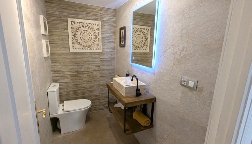 Baño contemporáneo con lavabo de piedra sobre encimera de madera, espejo con retroiluminación LED y paredes revestidas en tonos tierra con texturas naturales.