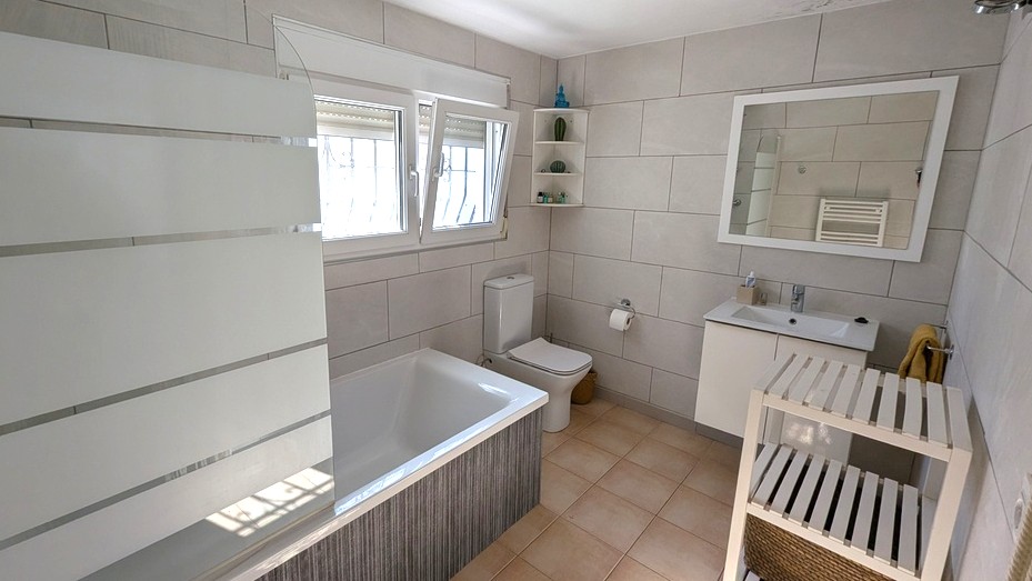Baño moderno con bañera integrada, mampara de cristal esmerilado, mueble lavabo blanco y paredes revestidas con azulejos de gran formato.
