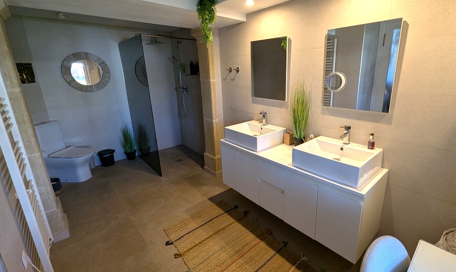 Baño moderno con lavabo de doble seno sobre mueble suspendido, ducha de obra con mampara oscurecida y acabados en tonos neutros con pilares de piedra.