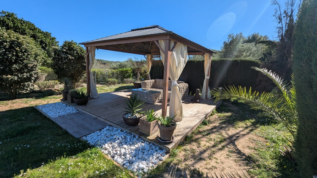 Cenador de madera sobre tarima exterior con mobiliario de mimbre, cortinas decorativas y zona de piedras blancas, rodeado de vegetación mediterránea en el jardín de Jávea.