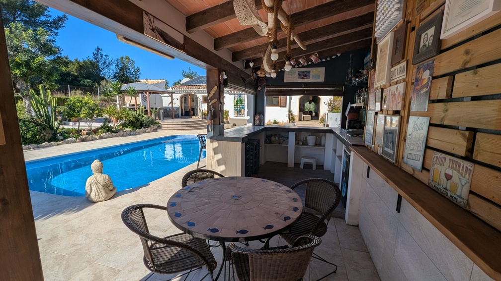 Cocina de verano y zona de bar con vigas de madera, mesa de mosaico y vistas a la piscina privada rodeada de vegetación mediterránea.