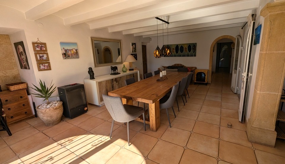 Comedor espacioso con una gran mesa de madera maciza, suelos de terracota y techos con vigas blancas. Un pilar de piedra Tosca y el arco de entrada realzan el carácter tradicional de la finca.