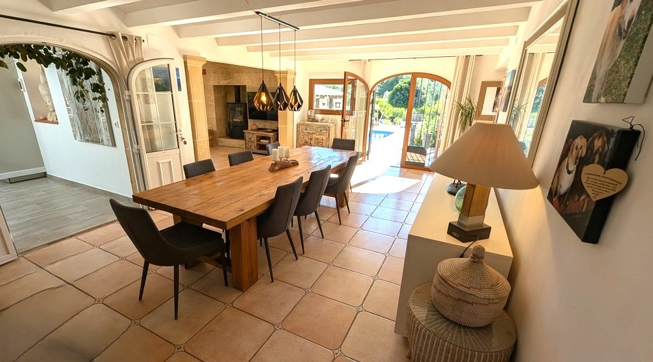 Comedor luminoso con mesa de madera maciza, vigas blancas vistas y suelo de terracota. Las puertas de arco acristaladas ofrecen acceso directo a la terraza y la piscina.