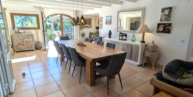 Comedor luminoso con mesa de madera rústica, vigas blancas y suelos de terracota, conectado al exterior mediante un gran ventanal de arco con vistas al jardín.