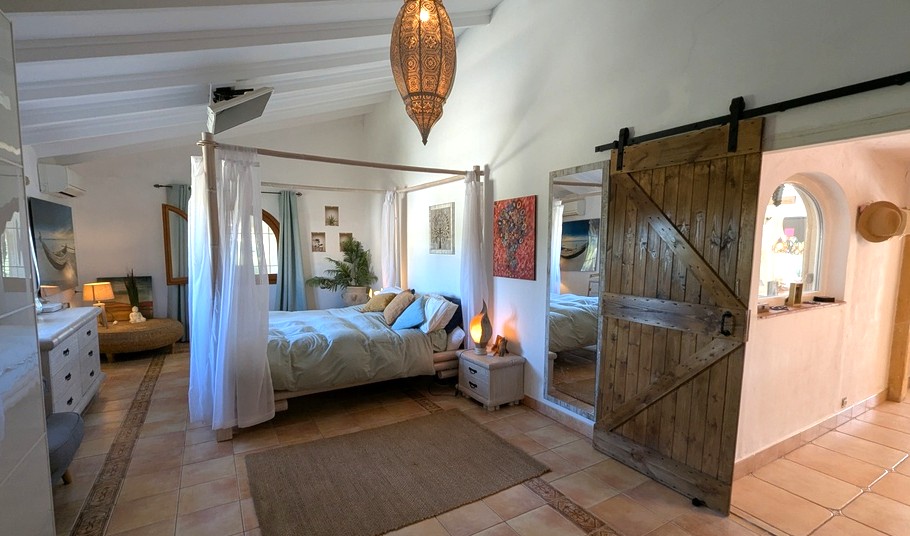 Dormitorio con cama de dosel, techos de vigas blancas y puerta corredera rústica. El suelo de terracota y la lámpara marroquí realzan el estilo mediterráneo.
