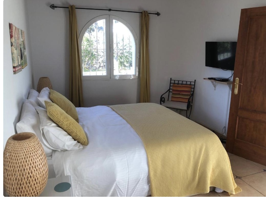 Dormitorio luminoso con ventana de arco, suelos de terracota y televisión de pared. La estancia destaca por sus detalles en tonos mostaza y lámparas de mimbre.