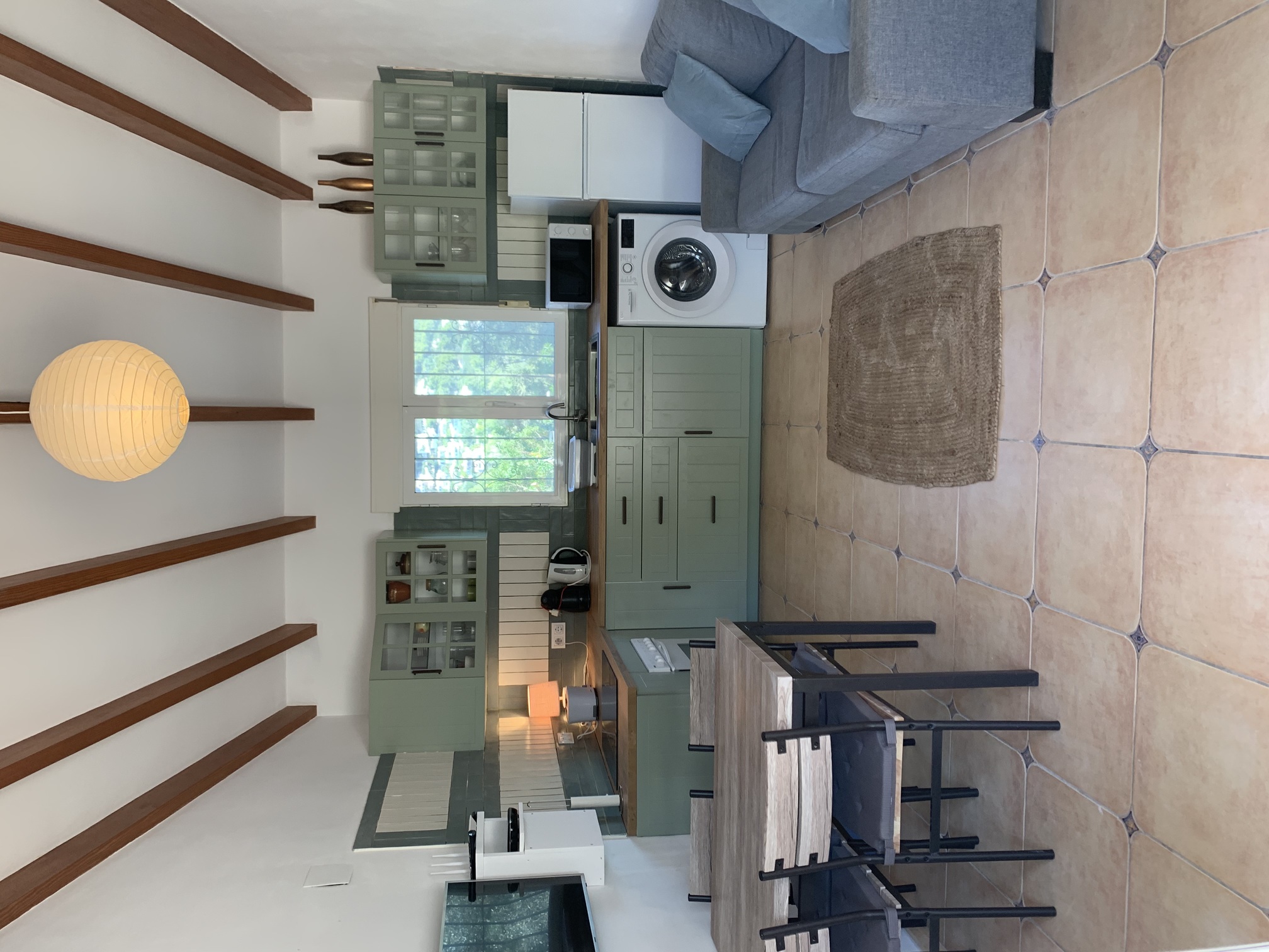 Espacio diáfano con cocina verde salvia, encimeras de madera, vigas vistas y zona de estar sobre suelos de gres tradicional con detalles decorativos.