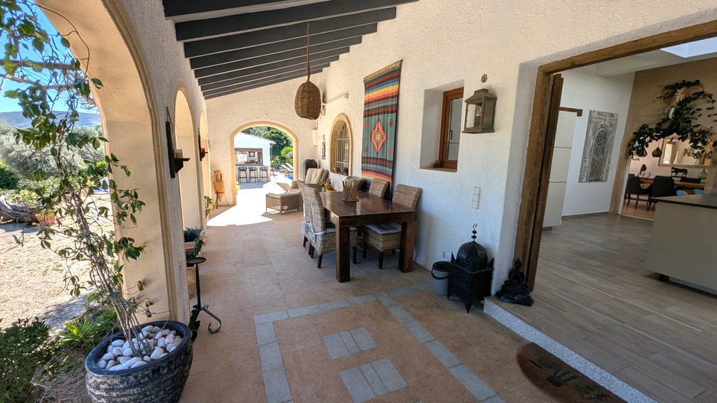 Naya tradicional con vigas de madera oscura, arcos de medio punto y zona de comedor. Esta terraza cubierta conecta el interior con el jardín y la cocina de verano.