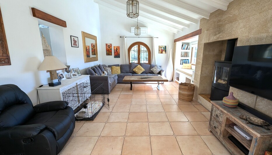 Salón espacioso con techos altos de vigas blancas, chimenea de piedra con estufa de leña y suelos de terracota. Incluye un ventanal de madera en arco y zona de lectura.