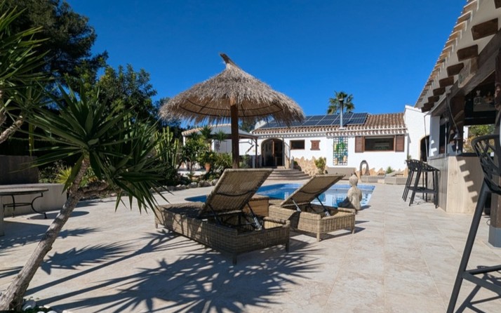 Terraza con piscina privada, tumbonas de mimbre bajo sombrilla de brezo y zona de bar. La vivienda de fondo muestra paneles solares y arquitectura tradicional mediterránea.