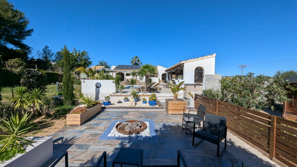 Terraza exterior con zona de hoguera central, pavimentos de pizarra y jardines escalonados que conducen a la villa principal con paneles solares.