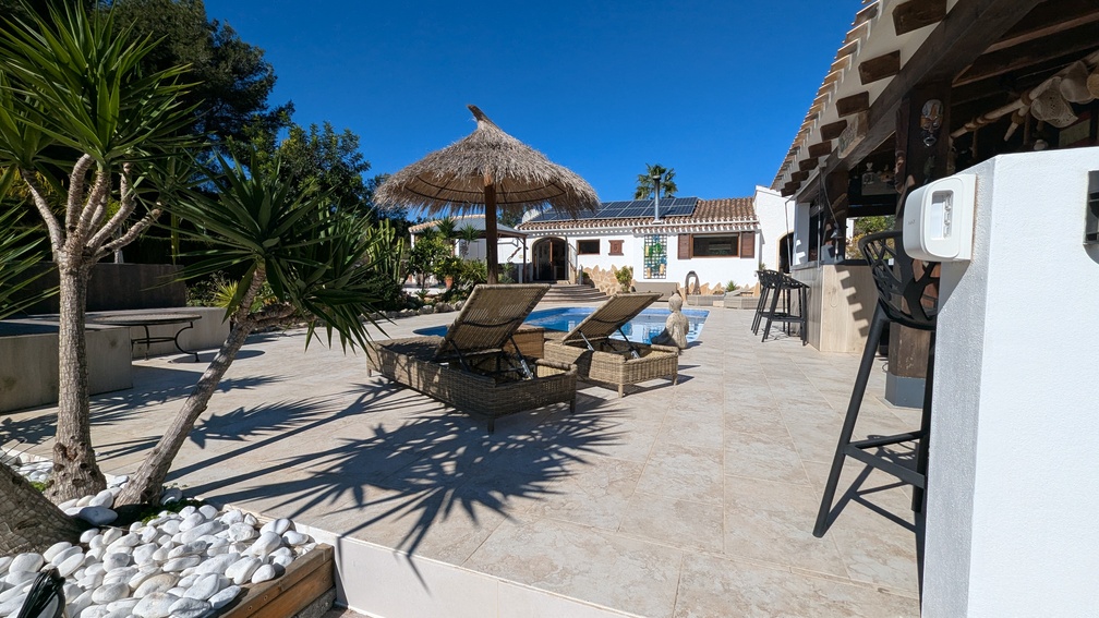 Terraza soleada con pavimentos cerámicos, tumbonas de mimbre y sombrilla de brezo junto a la piscina, con la villa principal y sus paneles solares al fondo.