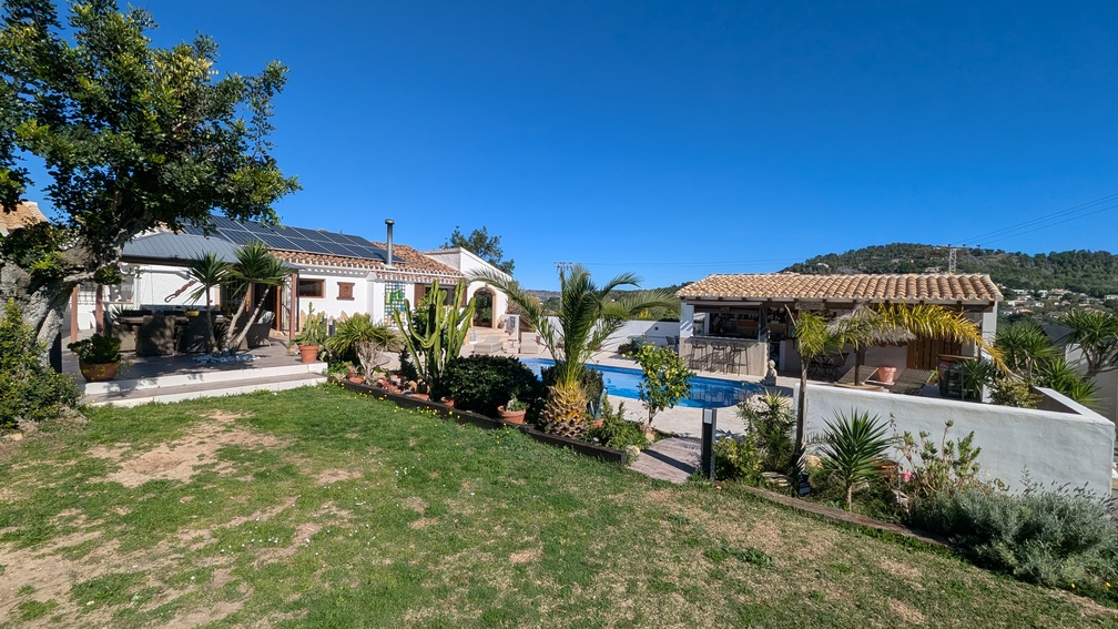 Vista exterior que muestra la casa principal con paneles solares, piscina privada y cocina de verano techada, rodeada de jardines y vegetación autóctona.